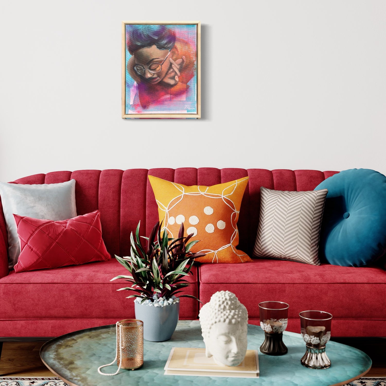 Ein modernes Wohnzimmer mit einem roten Samtsofa, das mit verschiedenen bunten Kissen, darunter orange, gelbe, blaue und weiße Muster, geschmückt ist. Eine Pflanze, Kerzen und eine Buddhakopfskulptur stehen auf einem runden Couchtisch. An der weißen Wand über dem Sofa hängt ein lebendiges Gemälde von Sabrina Wohlfeil Kunst.