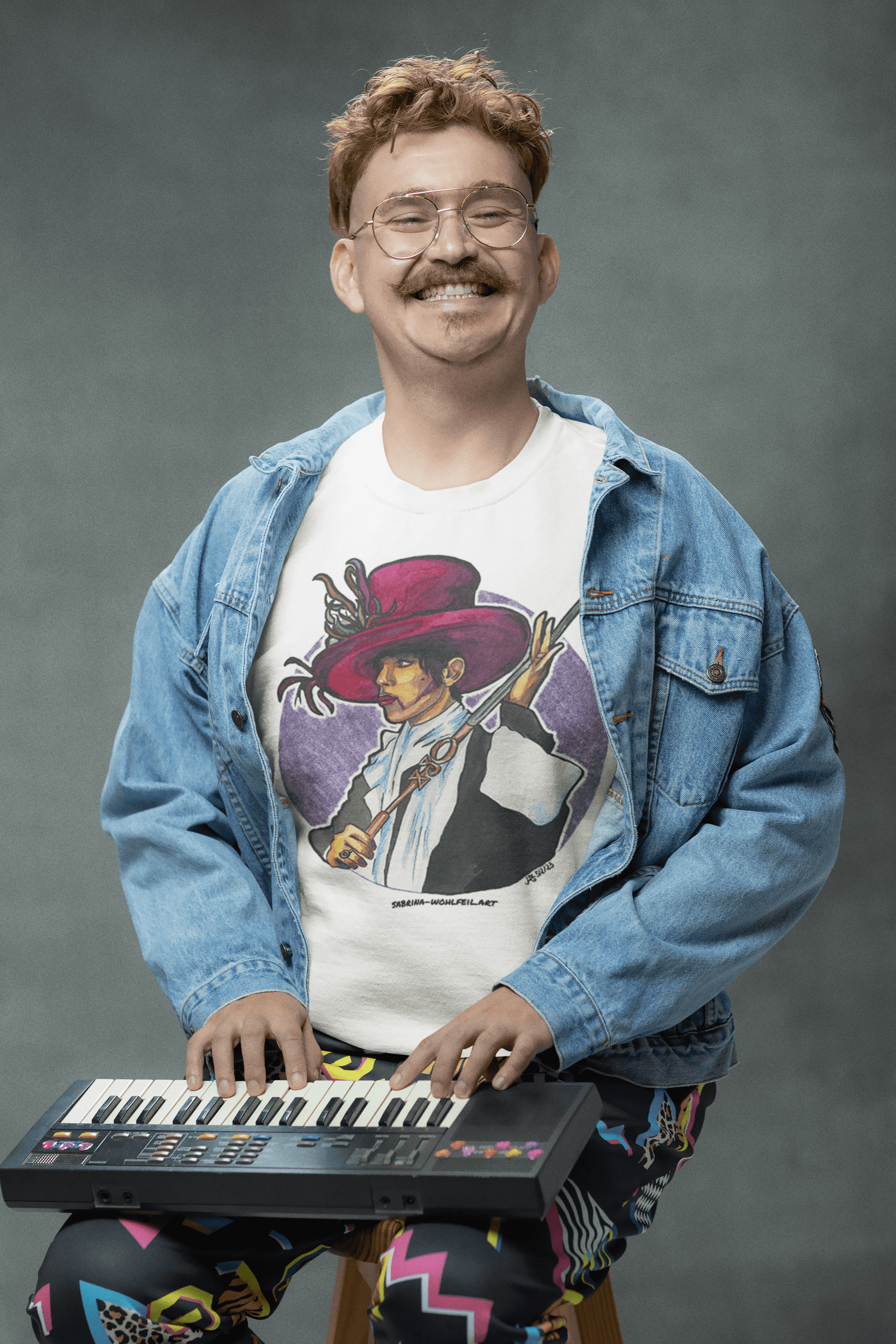 Smiling person wearing a denim jacket and a graphic t-shirt with an art print of Prince, playing a small keyboard.