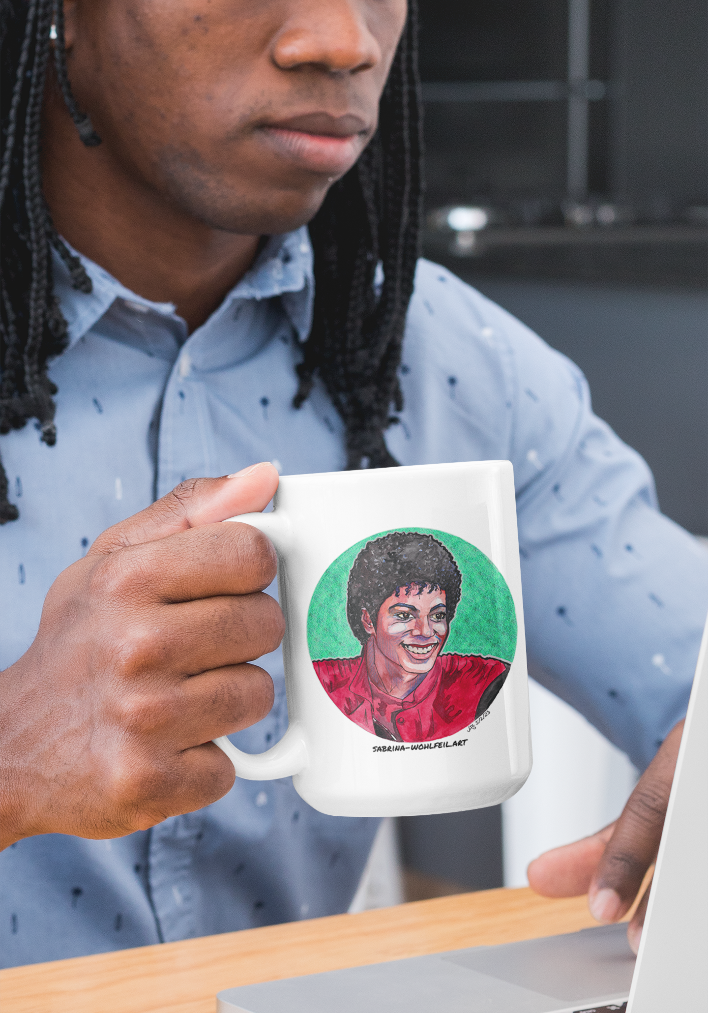 Mann hält weiße Keramiktasse mit Aquarellporträt von Michael Jackson, dem King of Pop, während er vor einem Laptop arbeitet.