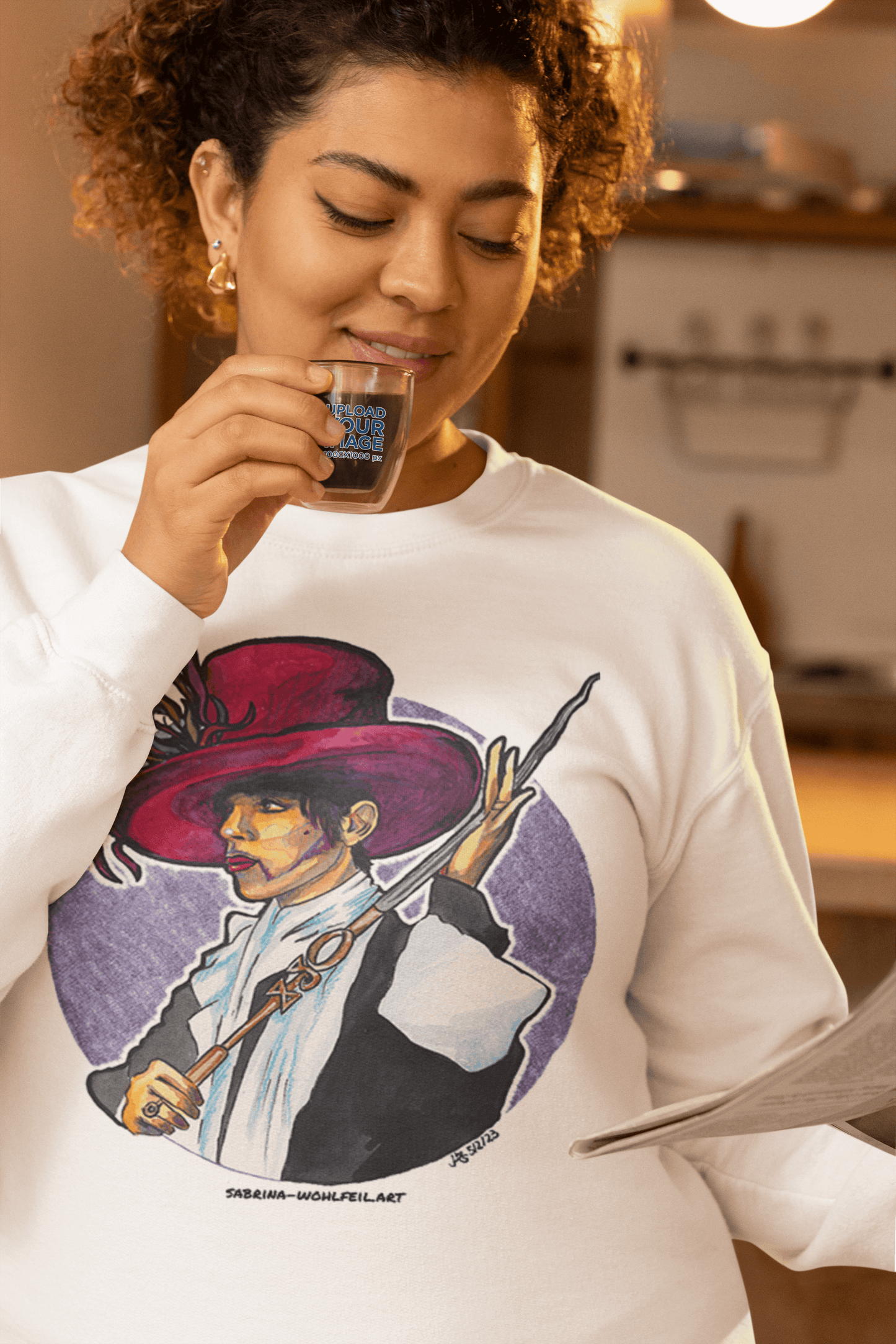 Person wearing a white unisex sweatshirt featuring a colorful Prince-inspired comic sketch by artist Sabrina Wohlfeil, holding a drink.